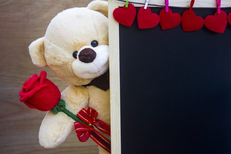 Valentine's day concept,Cute teddy Bear toy clutching a red rose in its arms and blackboard decorate red heart on wood background for an anniversary or valentine's celebration with copy space.の写真素材