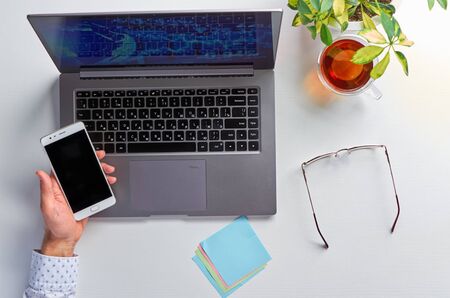 Male hands working on a laptop. Office workplace on a white table. In men's hands a mobile phone. The view from the top.の写真素材