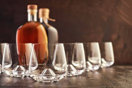 A set of five empty whiskey glasses on a wooden table. In the background are two closed bottles of whiskey.の写真素材