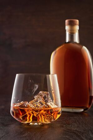 A crystal glass of whiskey on the rocks with a double portion on a wooden table. In the background is a full bottle of whiskey.の写真素材