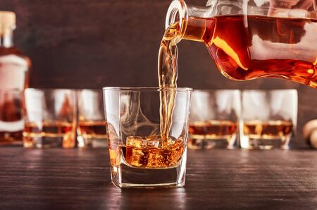 A glass of whiskey on a wooden table in which poured whiskey from a bottle. In the background are...