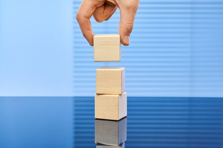 Three wooden cubes on a blue background standing on top of each other. A man's hand puts the cube on top. Free space for letters, numbers, symbols, or labels. Layout for infographics.の写真素材