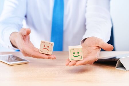 Business concept of satisfaction assessment, quality of service. The businessman holds in his hands a cube with drawings of sad and joyful emotions on each cube, respectively.の写真素材