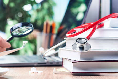 The stethoscope is lying on medical books, in the background a magnifying glass over a tablet in...