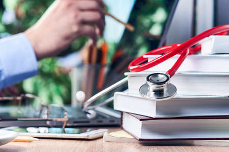 The stethoscope is lying on medical books, in the background a man's hand is holding a pencil and pointing it at the laptop screenの写真素材