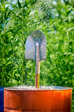 Garden shovel in a barrel of water in small splashes of water, against a background of green plants, place for textの写真素材