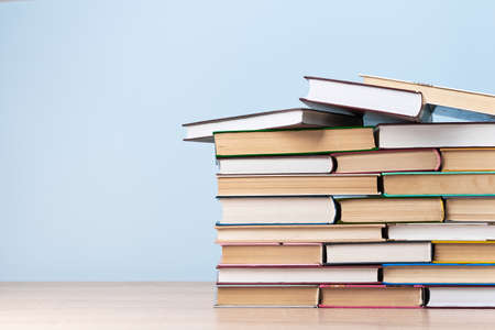 A large stack of books stands on a wooden table against a light blue wall, free space for text