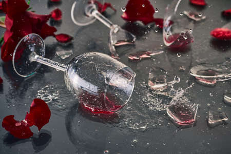 Two glasses of red wine, fallen and broken, lie on a glass table with glass fragments and petals of...