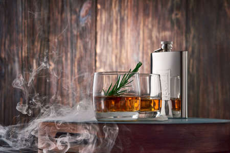 Two round glasses of whiskey with ice and a sprig of rosemary, and a metal flask, shrouded in clouds of smoke, stand on a tray against the old wooden wall. Low key.の写真素材