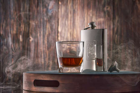 A square glass of whiskey and a metal flask stand on a tray against an old wooden wall, wreathed in clouds of smoke. Low key.の写真素材
