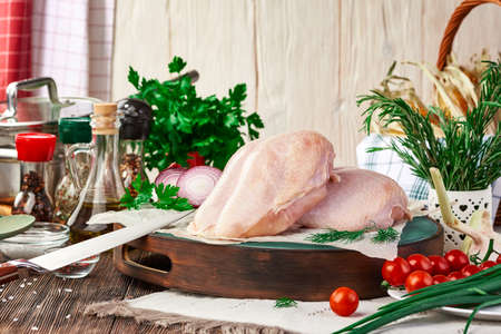 Two fresh raw chicken fillets on a cutting Board, on a dark wooden table with various kitchen utensils, spices, herbs and vegetables, cooking a chicken dish. Rustic still life.の写真素材