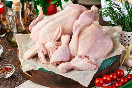 Chicken set of whole carcass, wings, leg and breast on a cutting Board, on a dark wooden table with various kitchen utensils, spices, herbs and vegetables, cooking chicken dishes. Rustic still lifeの写真素材
