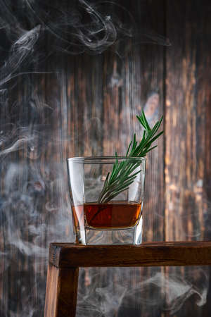 A square glass with a shot of whiskey and a sprig of rosemary on the counter with clouds of smoke, against the old wooden wall. Low key, vertical.の写真素材
