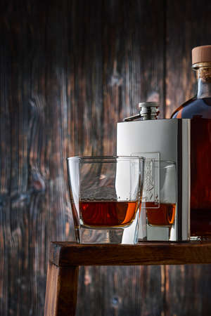 A square glass with a portion of whiskey, a metal flask and a full bottle of whiskey in a row on the counter, against the background of an old wooden wall. Low key, vertical.の写真素材