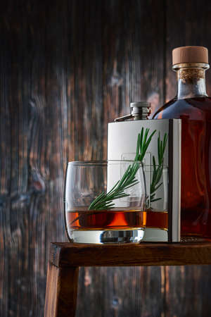 A round glass with a portion of whiskey and a sprig of rosemary, a metal flask and a full bottle of whiskey in a row on the counter, against the old wooden wall. Low key, vertical.の写真素材