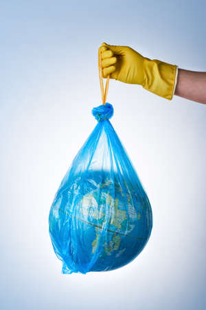 The concept of protecting the world from plastic waste, fighting environmental pollution, a hand in a yellow rubber glove holds a blue garbage bag with a globe on a light background. No plastic bagsの写真素材