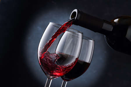 Red wine is poured into a glass from a bottle on a blurry blue background, a stream of red wine from the bottle swirls in the glass, close-up. Free space for text.の写真素材