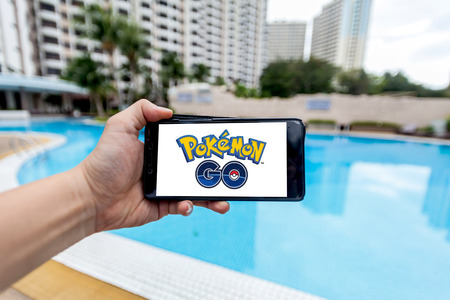 California, United States, 16 July 2016 : Hand holding a cellphone to play Pokemon Go with blur pool backgroundのeditorial素材