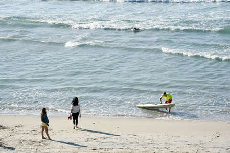 Young woman and girl seen rear view from top at seashore looking to the sea and children surfers near them on a sunny evening at Santa Comba beach, Ferrol, Galiza, Spainの写真素材