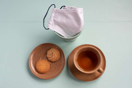 Knolling of a cup of tea, a couple of cookies and a crafted white facemask over light green aqua...