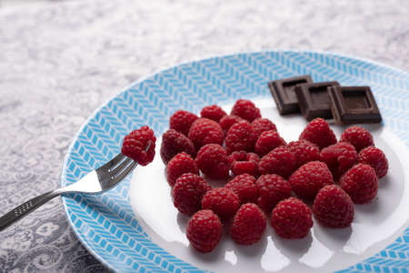 Fork with raspberry, heart of berries and chocolate squares on white and blue dish. Romantic...