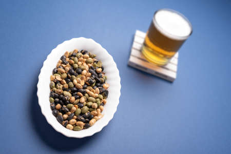 Snack mix of edamame and white and black toasted soy and blurred beer glass on coaster over a dark blue desk backgroundの写真素材