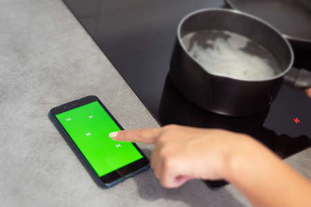Pointing finger to smartphone looking recipe next to boiling pot on kitchen stove in close-up....