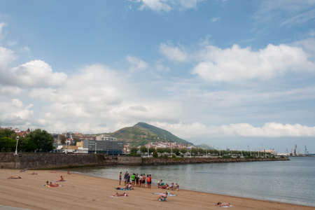 Getxo, Euskal Herria, Spain - 09 04 2018: People at Las Arenas beach and Serantes mountain on background in a sunny and cloudy day of summer. Churruca breakwater and shipyard cranes of Santurtziのeditorial素材