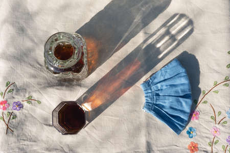 Tea glass and bottle and hygienic facemask flat lay at sunset on a linen tablecloth with floral pattern. Summer refreshing cold drink in pandemic with copy spaceの写真素材