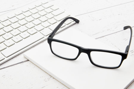 Black eyeglasses with keyboard on white wooden table, stock photo