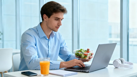 Handsome young man eating salad while working on laptop in officeの素材