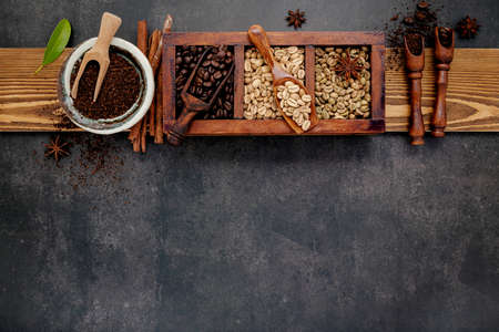 Green and brown unroasted and dark roasted coffee beans in wooden box with scoops setup on dark stone background.の写真素材