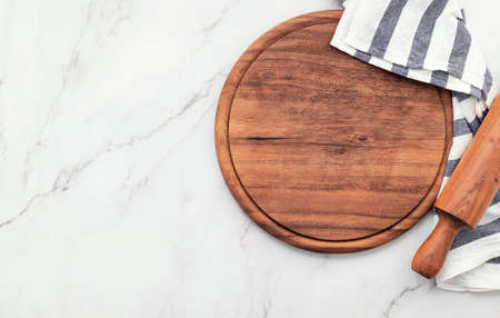Empty wooden pizza platter with napkin and rolling pin set up on marble stone kitchen table. Pizza board and tablecloth on white marble background.の写真素材