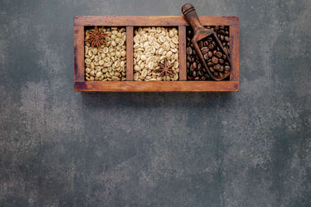 Green and brown unroasted and dark roasted coffee beans in wooden box with scoops setup on dark concrete background.の写真素材