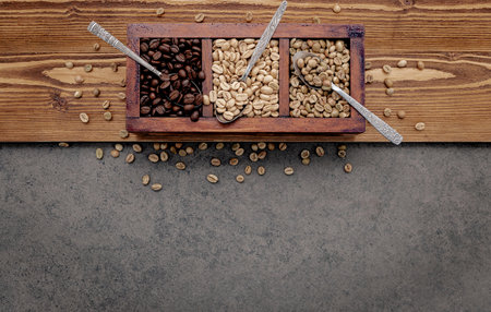 Green and brown unroasted and dark roasted coffee beans in wooden box with spoons setup on dark concrete background.の写真素材