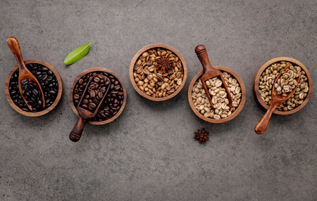 Green and brown unroasted and dark roasted coffee beans in wooden bowl with spoons set up on dark concrete background.の写真素材