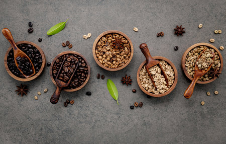 Green and brown unroasted and dark roasted coffee beans in wooden bowl with spoons set up on dark concrete background.の写真素材