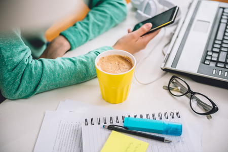 Yellow cup of coffee on the table by the laptop in the office in the background woman using smart phoneの写真素材
