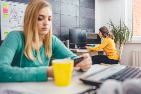 Woman working on the computer at the office and in the foreground girl sitting at the table and using smart phoneの写真素材