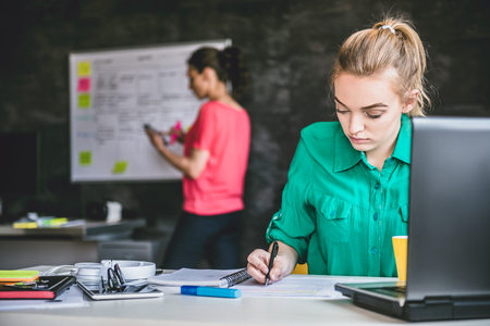 Young business woman wearing green shirt doing paperwork in the office and in the background is woman writing on whiteboardの写真素材