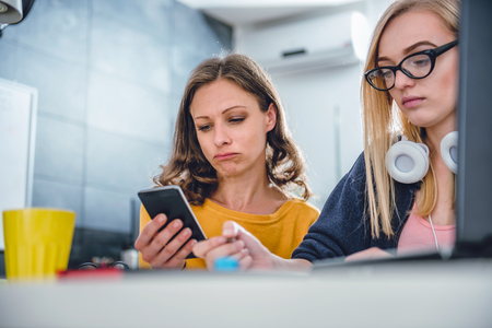 Two business woman working together at the office using smart phone and writing notesの写真素材