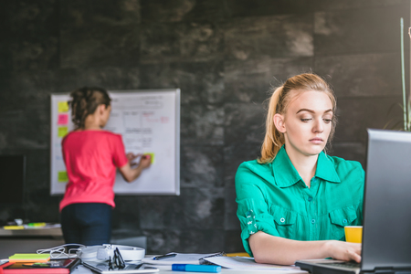 Young business woman wearing green shirt using laptop in the office and in the background is woman writing on whiteboardの写真素材