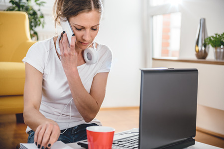 Woman wearing white shirt sitting at the desk and talking on the smart phone at homeの写真素材