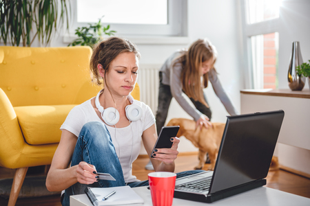 Woman wearing white shirt and headphones shopping online on smart phone and using credit card with a daughter and dog in the backgroundの写真素材