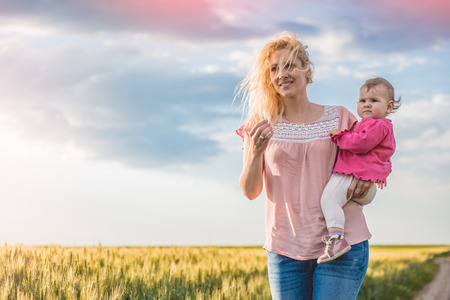 Mother holding her baby girl in her arms and walking on the gravel road by the wheat fieldsの写真素材