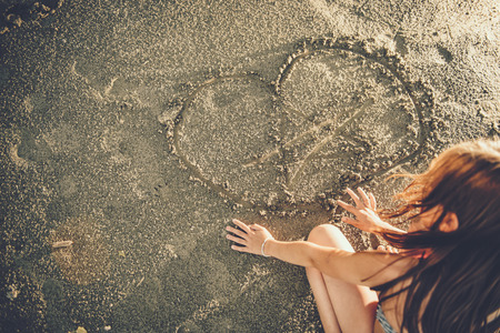 Girl drawing heart in the sand during sunsetの写真素材