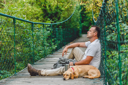 Hiker with small yellow dog resting on the wooden suspension bridge in the forestの写真素材