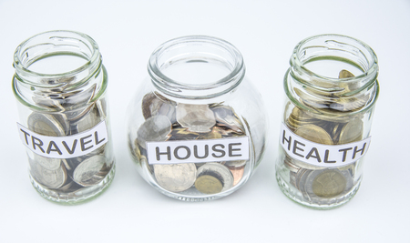 Coins in glass container with travel, house and health label. Selective focus.の写真素材