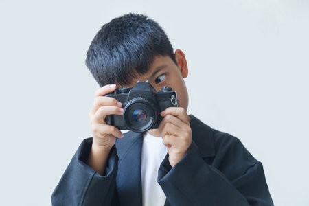A boy in a black suit holds an old film camera in his hand, against a white background.の写真素材