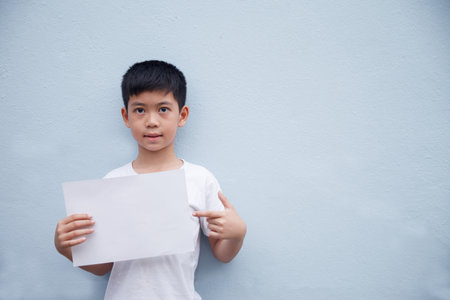 A boy wearing a white shirt holds a white piece of paper and points his finger at the white paper against a gray background.の写真素材